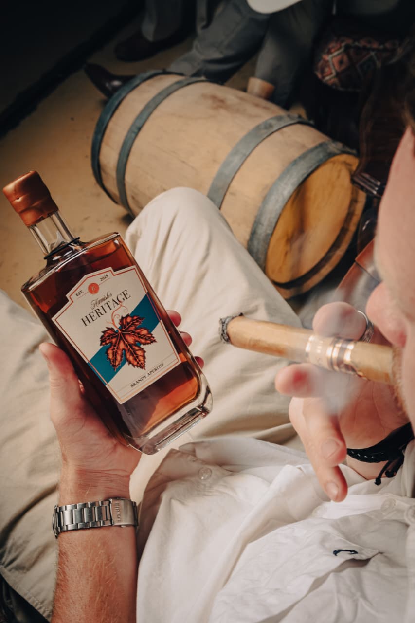Person holding a bottle of Heritage brandy and smoking a cigar with a wooden barrel in the background.